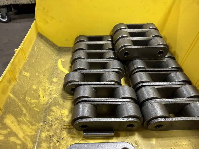 Stacks of machined conveyor chain links staged in a yellow shop bin.