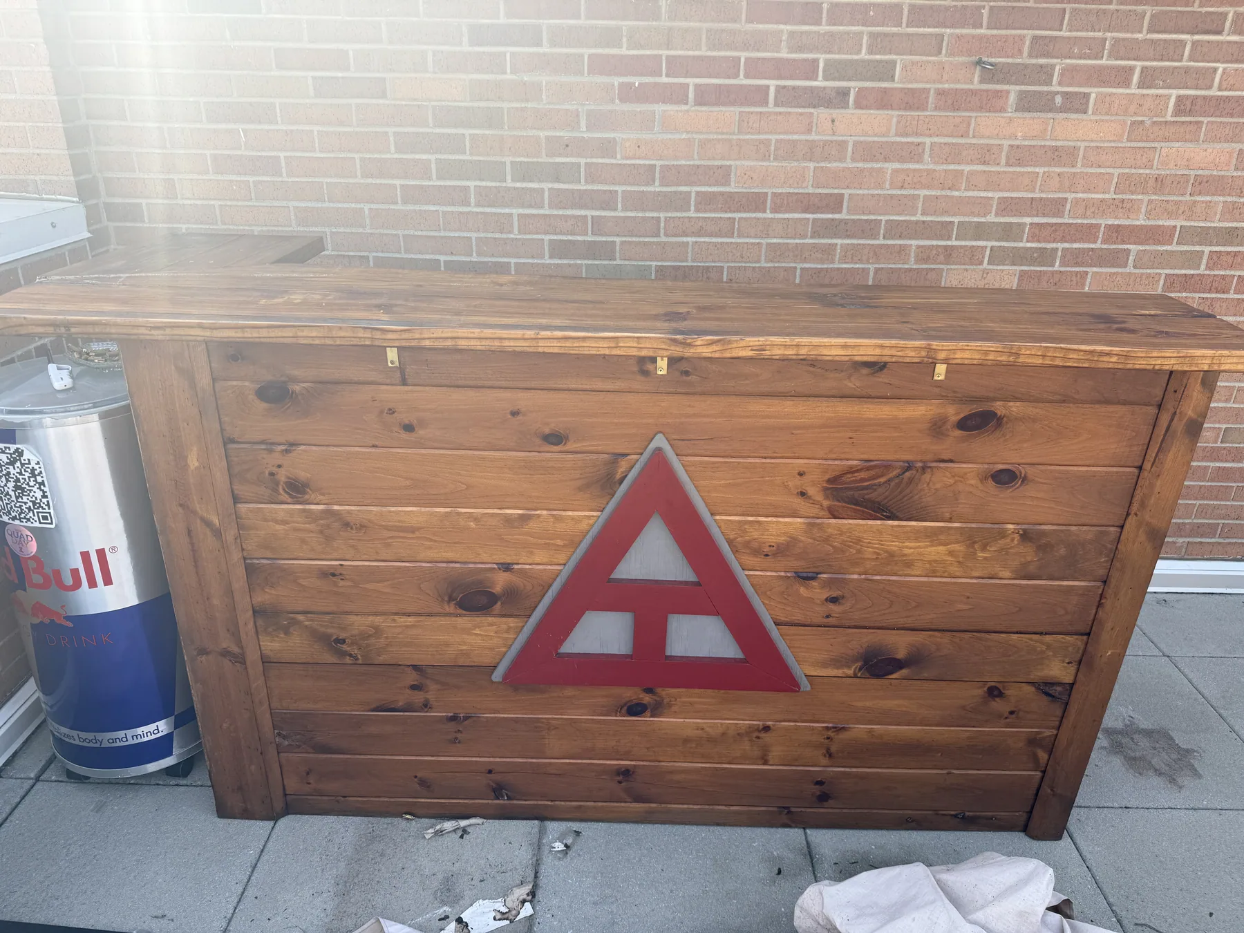 The finished structure seen from the front: a full-height stained timber bar with horizontal plank cladding, a thick overhanging top, and an inset painted triangular emblem.