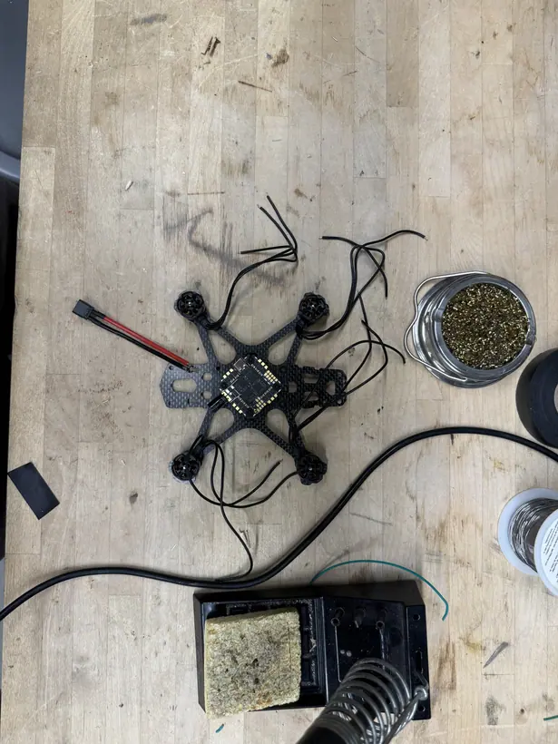 The partially built drone on a workbench beside a soldering station, tip cleaner, and solder spool: four motors mounted on the carbon-fiber frame, the electronics board at the center, and long unrouted motor leads spreading out across the bench.