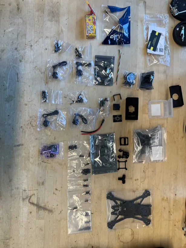 Overhead view of the drone components laid out on a workbench before assembly: the carbon-fiber frame plates, four bagged motors, the flight-control electronics in anti-static bags, the FPV camera and its mount, an antenna, a LiPo battery, a red and black power lead, and separate bags of fasteners.