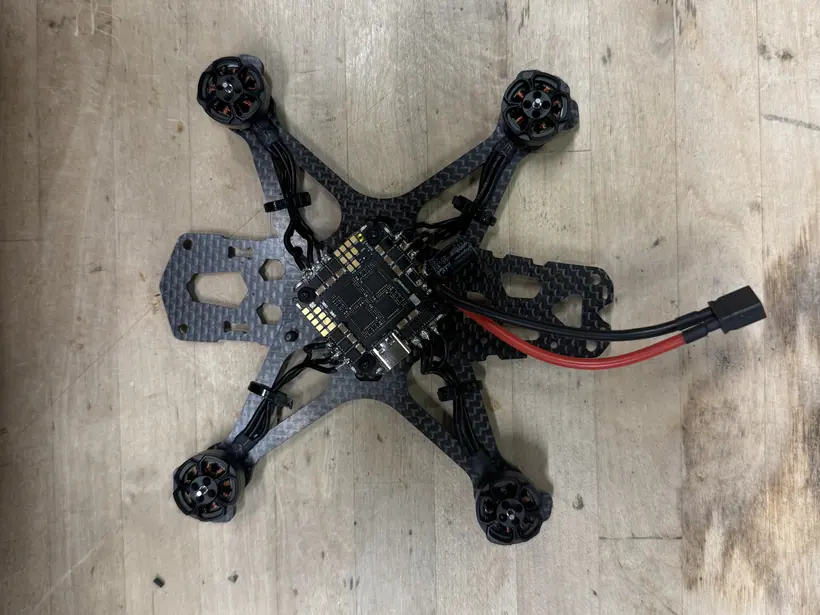 Current stage of the custom FPV drone: carbon-fiber quadcopter frame with four brushless motors, the flight-control board at the center, motor leads cable-tied along the arms, and the battery lead soldered in.