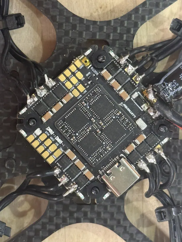 Close-up of the flight-control board mounted on the carbon-fiber frame, showing groups of three motor phase leads soldered to pads at each corner, rubber grommets on the mounting screws, the capacitor at the right edge, and cable ties holding the leads to the arms.